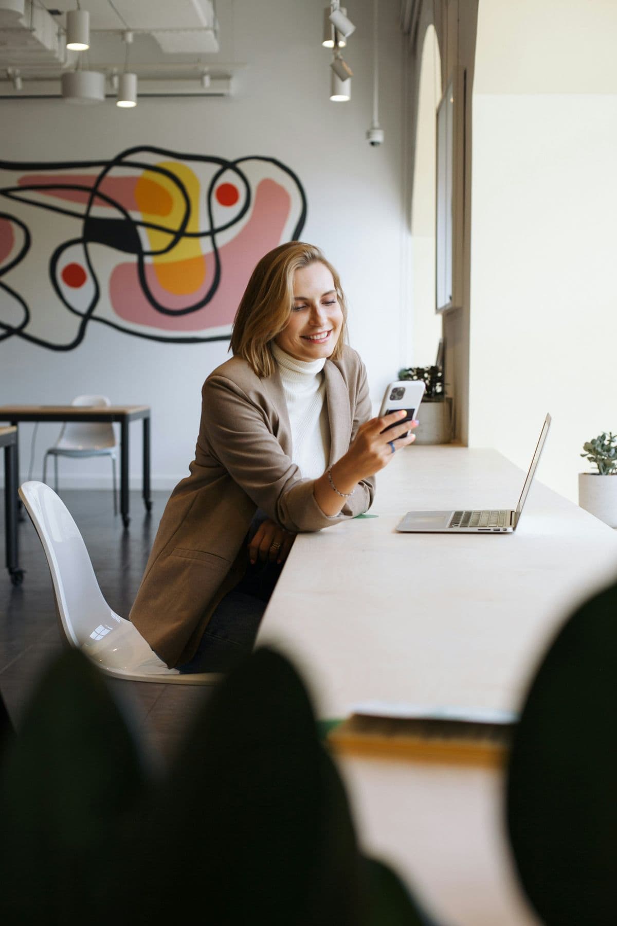 A professional checking her phone at a desk with a laptop in a bright modern office