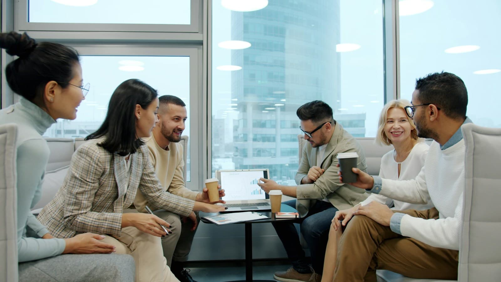 Colleagues in a bright glass-walled office meeting around laptops