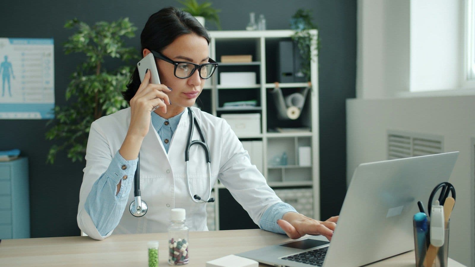 A doctor in a white coat taking a phone call while working at a laptop