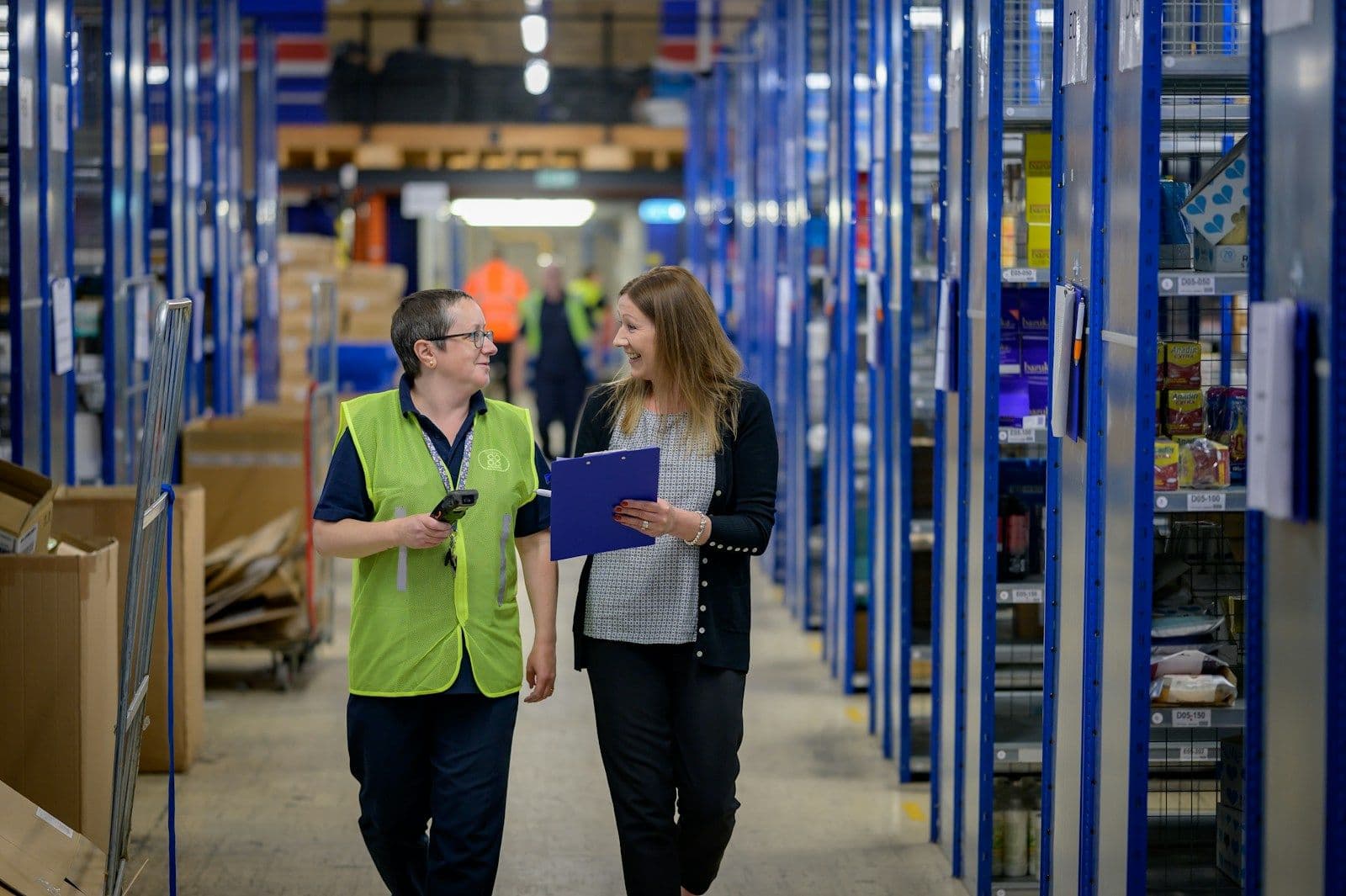 Two colleagues talking as they walk through a warehouse aisle, one holding a clipboard